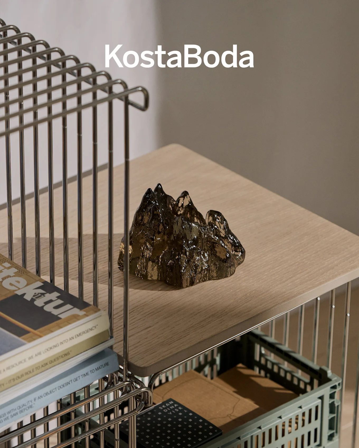 Nazwa marki KostaBoda nad ciemną, przezroczystą, górską szklaną rzeźbą na jasnej drewnianej półce.