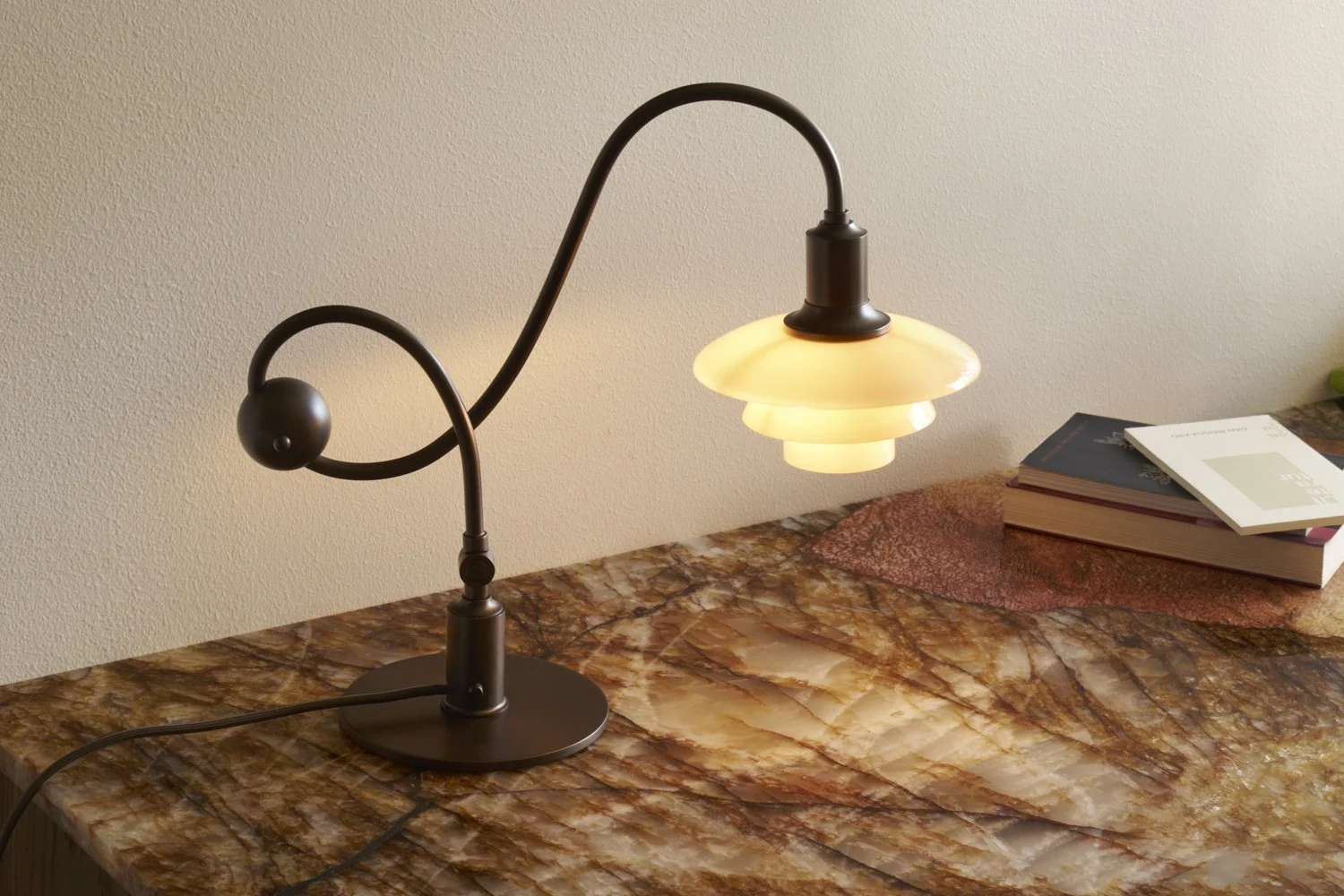Ciemna metalowa lampa stołowa z zakrzywioną podstawą i warstwowym, świecącym białym kloszem na marmurowej powierzchni z książkami.