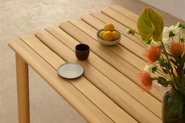 Jasny, drewniany stół lamelowy z miską cytryn, ciemnym kubkiem, małym talerzem i wazonem pomarańczowo-białych kwiatów.