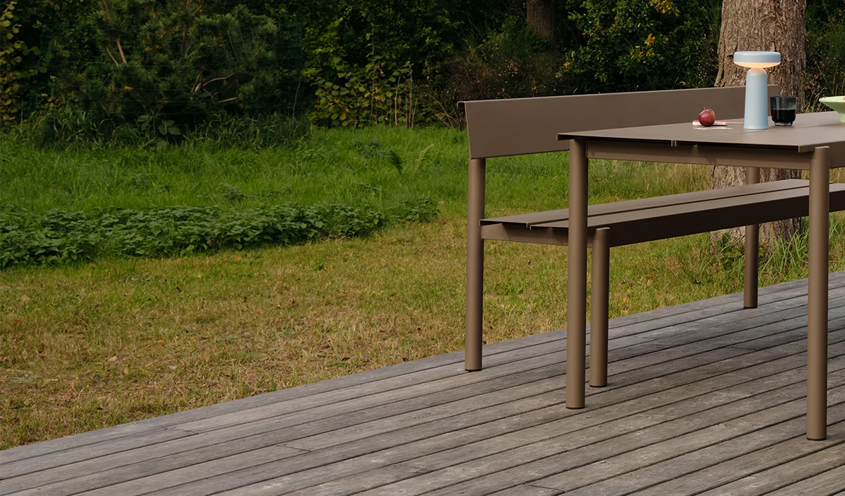 Nowoczesny stół i ławka jadalniana w kolorze taupe na drewnianym tarasie, z przenośną lampą i jabłkami na stole.