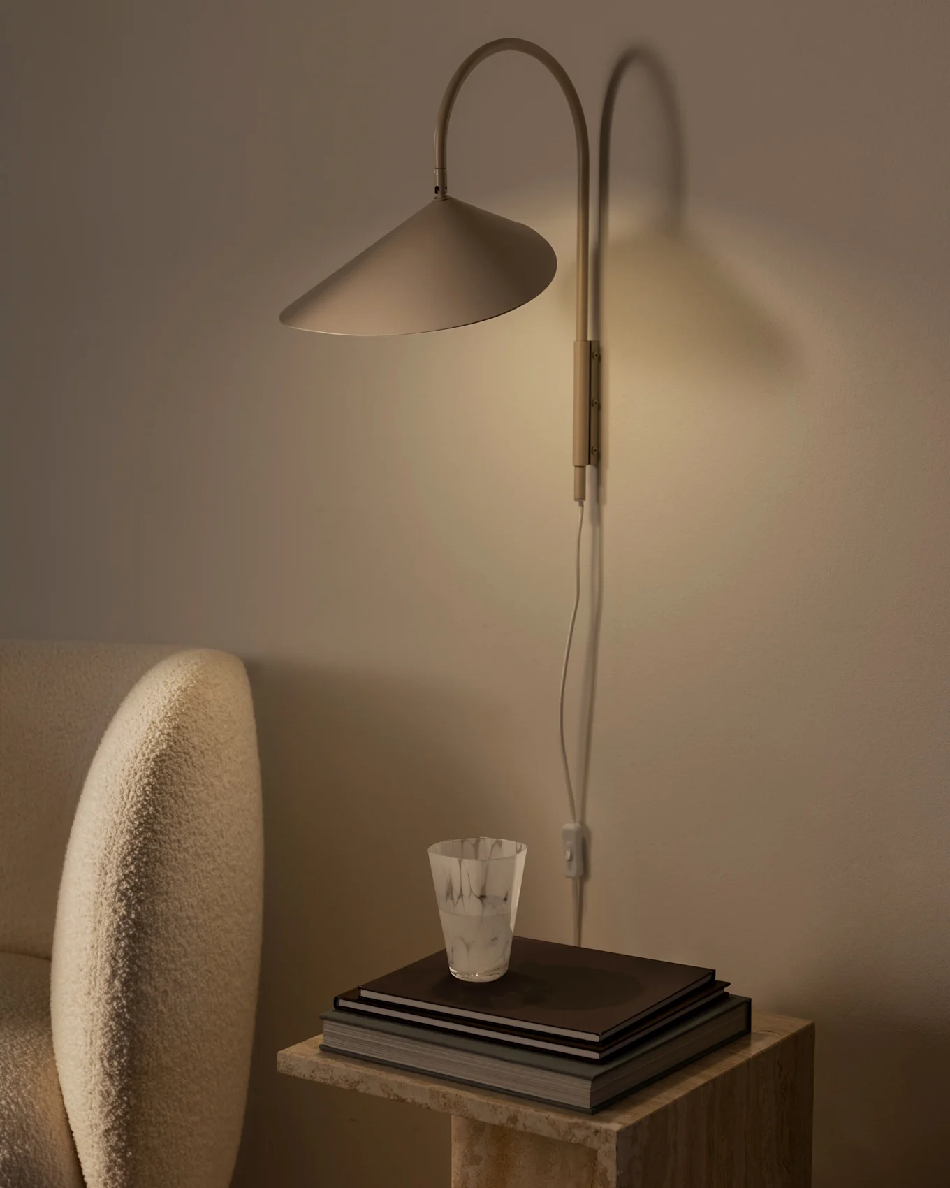 Ciepło oświetlona lampa ścienna z stożkowym kloszem nad stosem książek i teksturowaną szklanką na kamiennym stoliku, obok beżowego fotela.